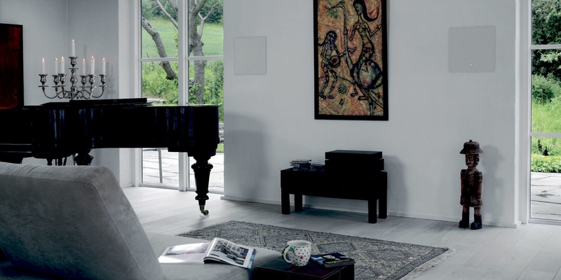 Two Dali H-60 in-wall speakers in wall with a white grille next to a black piano in a modern living space